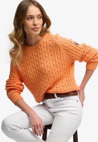 Orange kabelstrikket sweater med ribbede manchetter og kant. Båret over hvide bukser, der har et vævet brunt bælte. Indeholder et lille mærke på ærmet.