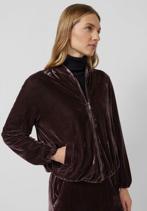 Vrouw in een donkerpaars fluwelen zip-up jack over een zwarte coltrui, staande met een hand in de zak, kijkend naar rechts tegen een effen achtergrond.