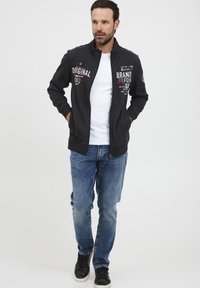 Mørkegrå zip-up jakke med broderte tekstdetaljer, brukt over en hvit skjorte. Kombinert med blå denimjeans og svarte joggesko.