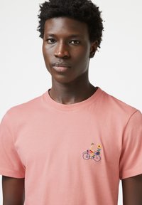 Roze katoenen T-shirt met een klein geborduurd ontwerp van een persoon op een fiets met een zonnebloem in een mand op de linkerborst.