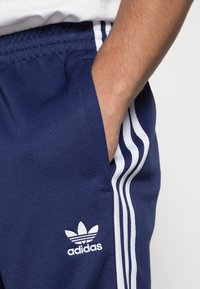 Námořnické modré tepláky Adidas s bílým designem tří pruhů po stranách, kartáčovanou strukturou a malým bílým logem na levém stehně.