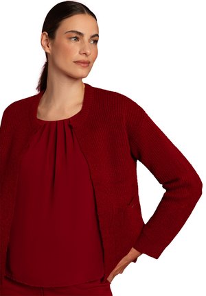 Roter Strickcardigan mit rundem Ausschnitt, strukturiertem Muster und offenem Vorderteil. Getragen über einem passenden roten plissierten Oberteil.