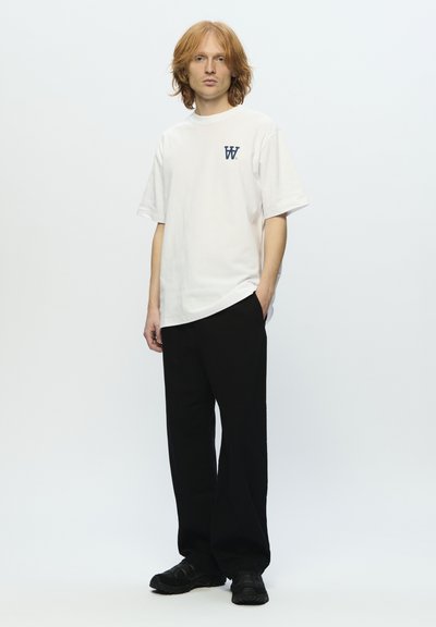 T-shirt bianco in cotone con un piccolo logo navy sul petto sinistro, abbinato a pantaloni neri larghi e sneaker scure.