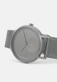 Grå nätarmbandsur med en rund grå urtavla, silverfärgade tim- och minutvisare samt en orange sekundvisare. Varumärket visas på urtavlan.