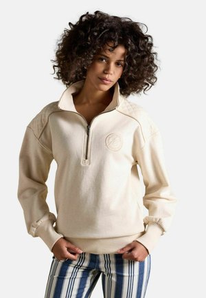 Frau mit lockigem Haar, die einen cremefarbenen Reißverschluss-Sweatshirt und blau-weiß gestreifte Hosen trägt, steht mit den Händen in den Taschen.