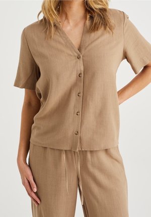 Femme portant un haut beige à manches courtes avec boutons et un pantalon ample assorti, debout avec une main sur la hanche et l'autre détendue.