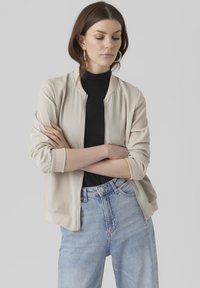 Light beige bomber jacket with a ribbed collar and cuffs, zip front, and soft texture, paired with high-waisted light blue jeans.