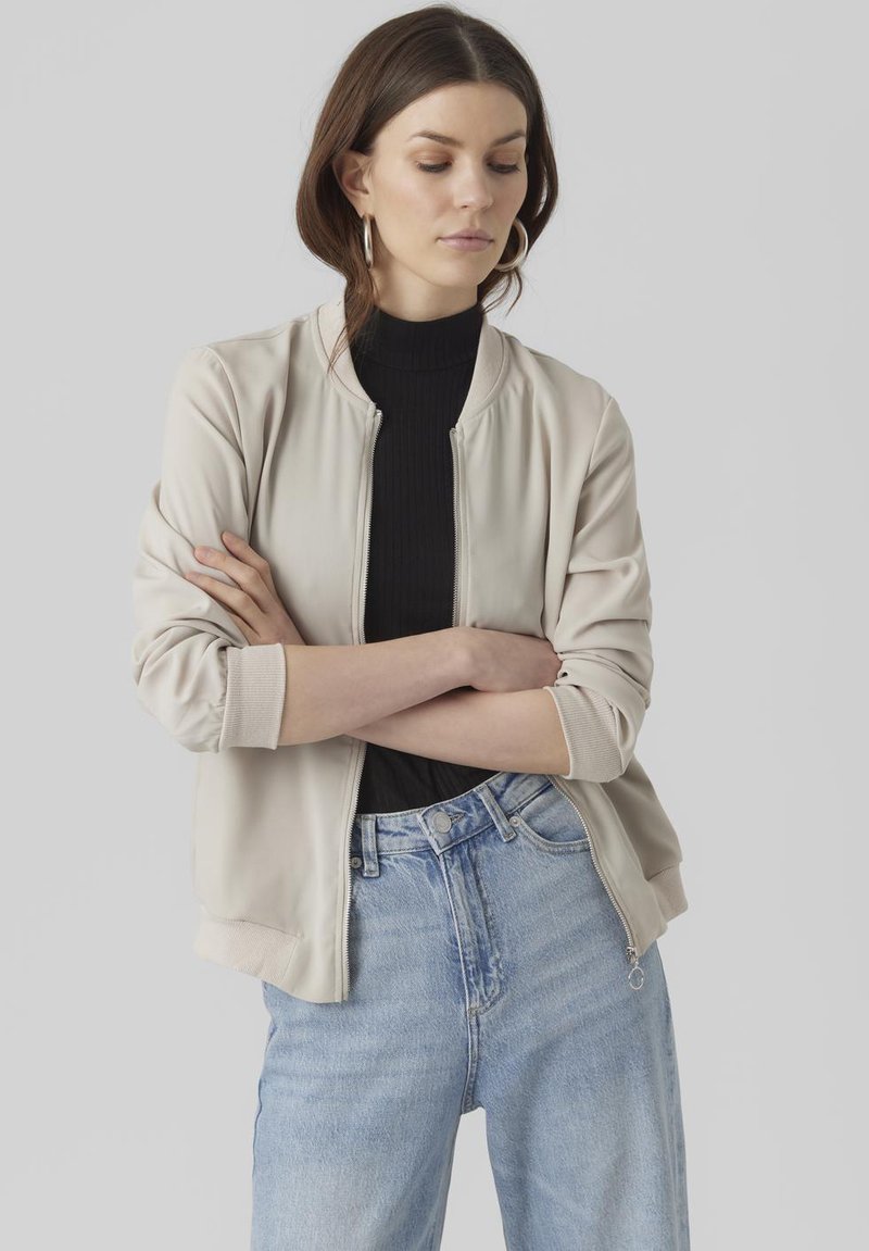 Light beige bomber jacket with a ribbed collar and cuffs, zip front, and soft texture, paired with high-waisted light blue jeans.