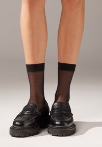 Medias tobilleras negras traslúcidas combinadas con mocasines de cuero negro con suela gruesa y un diseño minimalista. Textura suave y ajuste ceñido.