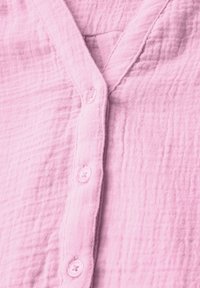 Camicia button-up di colore rosa chiaro con un tessuto texturizzato e stropicciato, scollatura a V e quattro bottoni coordinati sulla parte anteriore.