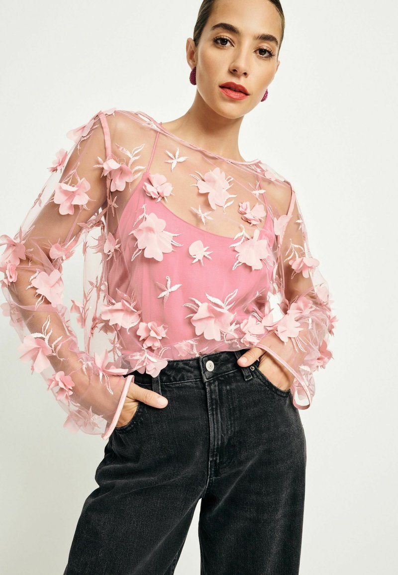 Genomskinlig rosa blus med blommönster och långa ärmar, lager på lager över en enfärgad rosa innertröja, kombinerad med högmidjade svarta jeans.