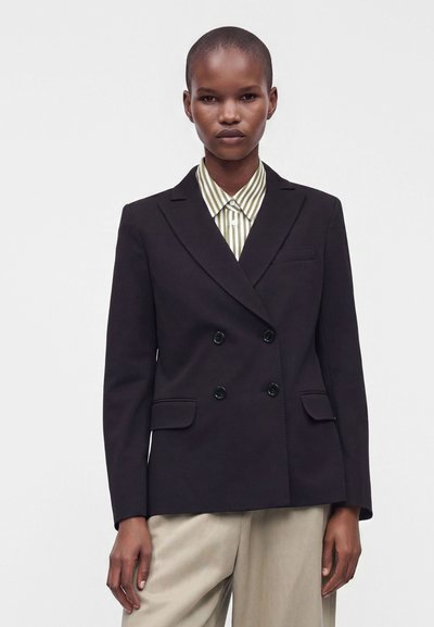 Personne aux cheveux courts portant un blazer noir à double boutonnage sur une chemise à col rayée vert et blanc et un pantalon beige.