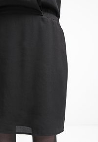 Jupe noire longueur genou portée sur des collants noirs transparents, vue de la taille au milieu de la cuisse sur un fond blanc.