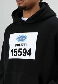 Zwarte hoodie met een voorzak, een witte rechthoekige patch met de tekst "POLIZEI" en "15594" in vet zwarte letters, en een blauw logo erboven.