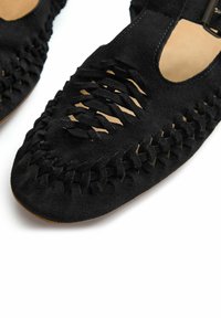 Zapatos de ante negros con lados abiertos, que presentan un cordón intrincado y un diseño trenzado a lo largo de los bordes. Punta redondeada y plana con un interior suave.