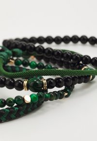 Többszínű gyöngy bracelet, amely fekete és zöld gyöngyökből áll, zöld fonott zsinórral és arany díszítéssel össze intertwined.