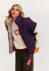 Puchowa fioletowa kurtka z podszewką w panterkę, zestawiona z kremowym swetrem o teksturze, ozdobionym kolorową haftowaną aplikacją oraz brązowymi spodniami w panterkę.
