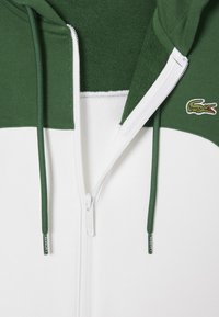 Groene en witte zip-up hoodie met een textuur op het bovenste deel, een gladde onderkant, een trekkoord en een klein geborduurd krokodillenlogo.