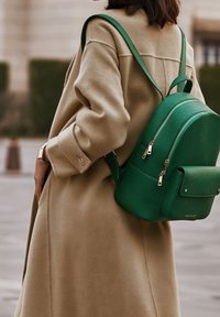Grön läderryggsäck med två dragkedjefack och en framficka, bärs över en beige kappa med en texturerad yta. Guldhardware detaljer.