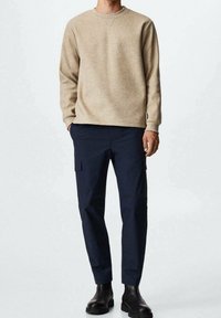 Beżowy sweter z fakturowanej tkaniny, z okrągłym dekoltem i długimi rękawami, w połączeniu z granatowymi spodniami typu cargo z dwoma kieszeniami po bokach i zwężanym krojem. Czarne buty.