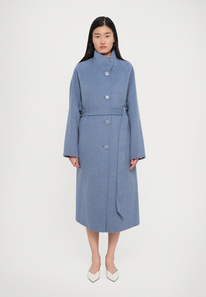 Femme portant un manteau bleu mi-long ceinturé avec de grands boutons et un col montant, associé à des chaussures blanches à bout pointu sur un fond uni.