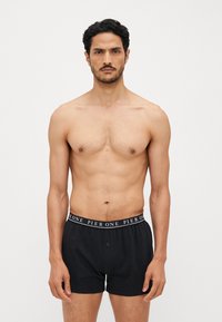 Boxers en coton noir avec fermeture à bouton et une ceinture élastique à motifs affichant "PIER ONE." Le modèle se tient devant un fond uni.