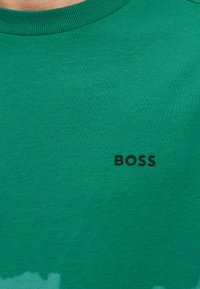 Πράσινο πουλόβερ με στρογγυλή λαιμόκοψη και μαύρο λογότυπο "BOSS" τυπωμένο στο στήθος, που φοράει ένα άτομο.