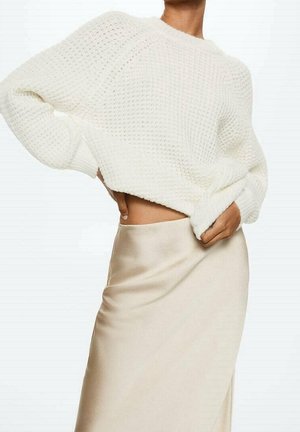 Femme portant un gros pull en maille blanc et une jupe en satin lisse, couleur crème, une main sur la hanche et l'autre ajustant le pull.