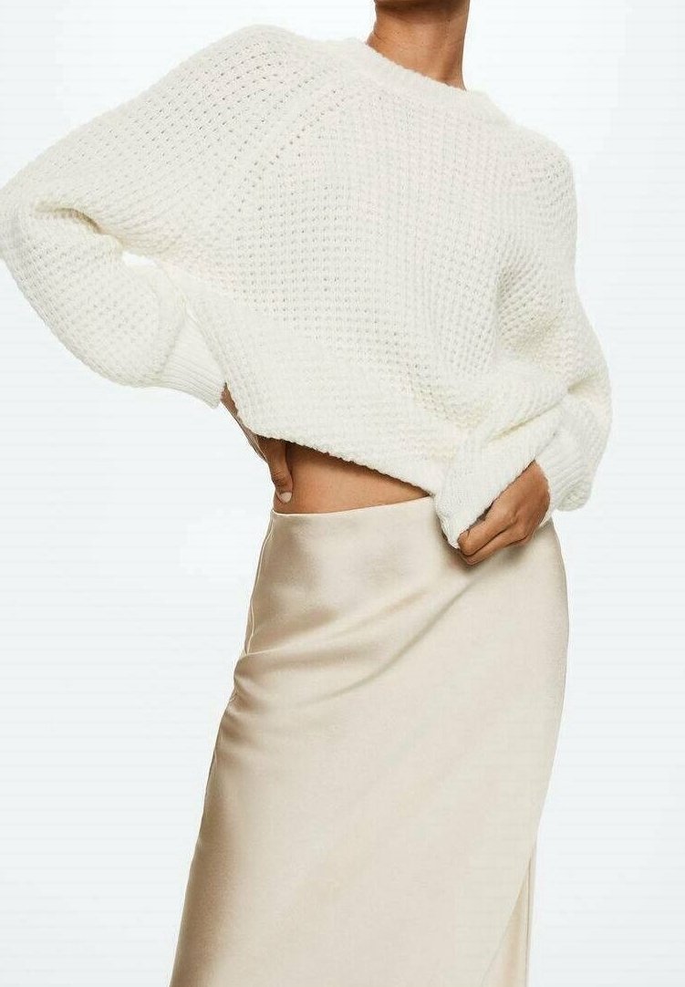 Femme portant un gros pull en maille blanc et une jupe en satin lisse, couleur crème, une main sur la hanche et l'autre ajustant le pull.