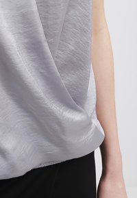 Haut sans manches en satin gris clair porté sur le torse d'une personne, montrant un tissu drapé et une partie du bras droit sur un fond uni.
