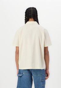 Camisa de manga corta color crema claro con cuello, corte recto, textura sutil y una costura en la parte posterior, combinada con pantalones cortos de mezclilla tipo cargo.