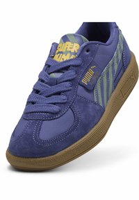 Violet sporta apavi, kas sastāv no gludas ādas un zamšāda, ar zaļiem svītru akcentiem un brūnu gumijas zoli. Auklu dizains ar logotipa detaļām.