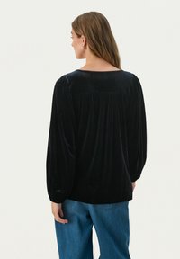 Zwarte fluweel blouse met lange, opgeblazen mouwen, een brede halslijn en geplooide details op de achterkant, gecombineerd met blauwe denimbroek.