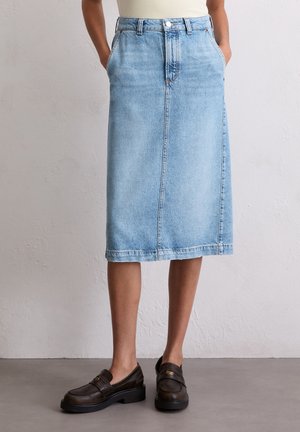 Mujer con una falda vaquera azul claro hasta la rodilla con bolsillos y mocasines de cuero marrón, de pie sobre un suelo gris.