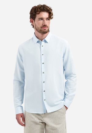 Man met krullend haar en baard draagt een lichtblauw overhemd met knopen en beige broek met dunne verticale zwarte strepen, staand met één hand in de zak.