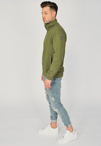 Zielona oliwkowa kurtka na zamek z wysokim kołnierzem, teksturowana tkanina, kieszenie boczne i logo na lewym rękawie, zestawiona z jasnoniebieskimi dżinsami w stylu distressed i białymi sneakersami.