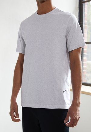 Mand iført almindelig lys grå t-shirt med korte ærmer og lille sort logo nær kanten, stående indendørs ved vindue med murstens- og hvide vægge.