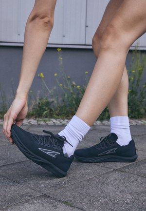 Črne športne čevlje z teksturiranim mrežastim zgornjim delom, vezalkami in odsevni akcenti, v kombinaciji z belimi nogavicami do gležnjev, na asfaltirani površini.