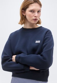 Νέα γυναίκα με κοντά ξανθά μαλλιά, που φοράει χρυσούς κρίκους και μια σκούρα μπλε μπλούζα με μικρό λευκό λογότυπο, με τα χέρια σταυρωμένα.