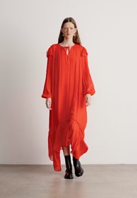 Largo vestido rojo hecho de tejido ligero con un escote alto, mangas acampanadas y un dobladillo asimétrico, combinado con botas negras hasta el tobillo.