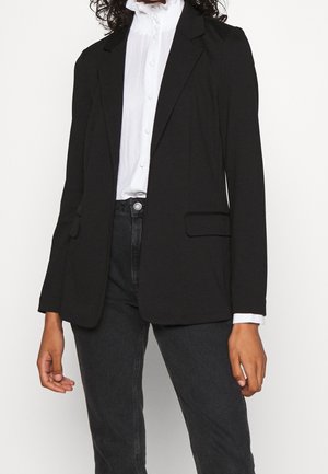 Persoon die een zwarte op maat gemaakte blazer draagt over een wit gebuttoned overhemd met geplooide kraag en zwarte spijkerbroek, staand tegen een effen achtergrond.