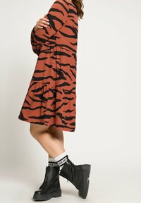 Sukienka w kolorze rdzy z czarnymi tygrysimi paskami, warstwowa konstrukcja, długie rękawy, w połączeniu z czarnymi botkami za kostkę i białymi skarpetkami z napisem.