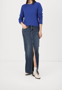 Pull bleu à manches longues avec des poignets côtelés, associé à une jupe en jean taille haute avec une fente à l'avant, et des chaussures plates beiges claires.