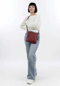 Rote Leder-Crossbody-Tasche mit einem Reißverschlussfach, glatter Textur und verstellbarem Riemen. Kombiniert mit hellblauen Schlagjeans und einem weißen Pullover.