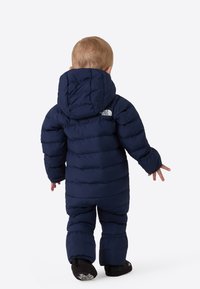 Mørkeblå boblejakke og -bukse med hette, med quiltet tekstur og en logo på baksiden. Myke tøfler fullfører antrekket.