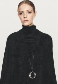 Kvinna med ljus hy och hår uppsatt bär en fluffig svart polotröja och ett långt halsband med en metallisk oval berlock.