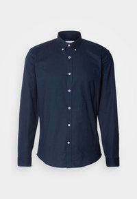 Chemise bleu marine à manches longues avec boutons blancs et col boutonné, présentée sur un fond blanc uni.