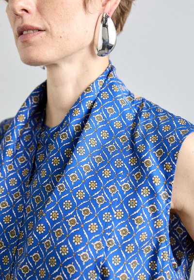 Blau gemusterter Stoffoberteil mit floralen Designs in gelben und weißen Akzenten. Silberne Tropfenohrringe fügen ein metallisches Detail hinzu.