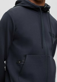 Mörkgrå zip-up hoodie av mjukt material, med en känguruficka och dragsko på huvan med accentuerad hårdvara.