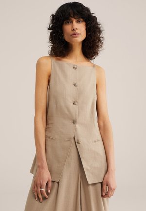 Žena s kučeravými tmavými vlasmi oblečená v béžovom bezrukávovom topu na gombíky a zodpovedajúcich širokých nohaviciach na jednoduchom pozadí.